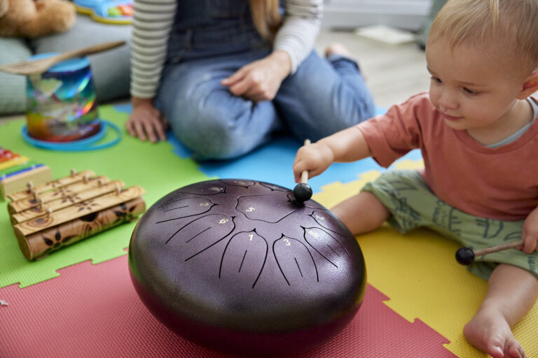 Toddler playing with musical instrument in playroom with mother sitting nearby